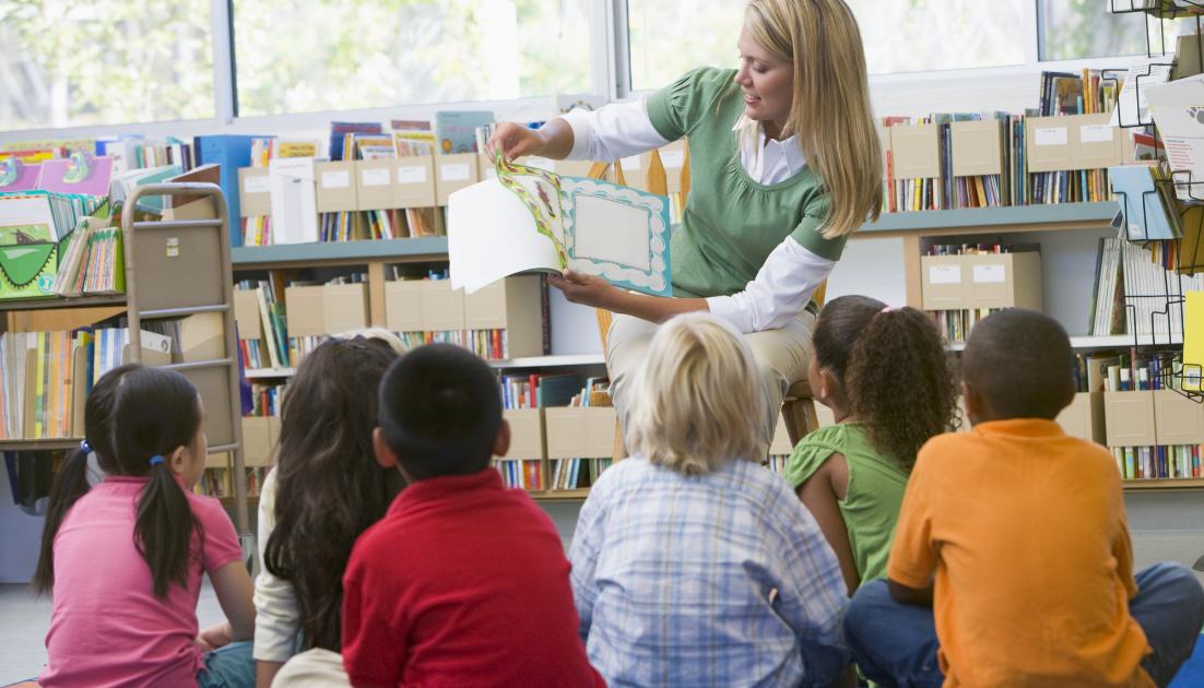 leggere-per-crescere Volontaria seduta di fronte a un gruppo di bambini seduti di spalle, che legge e mostra un libro